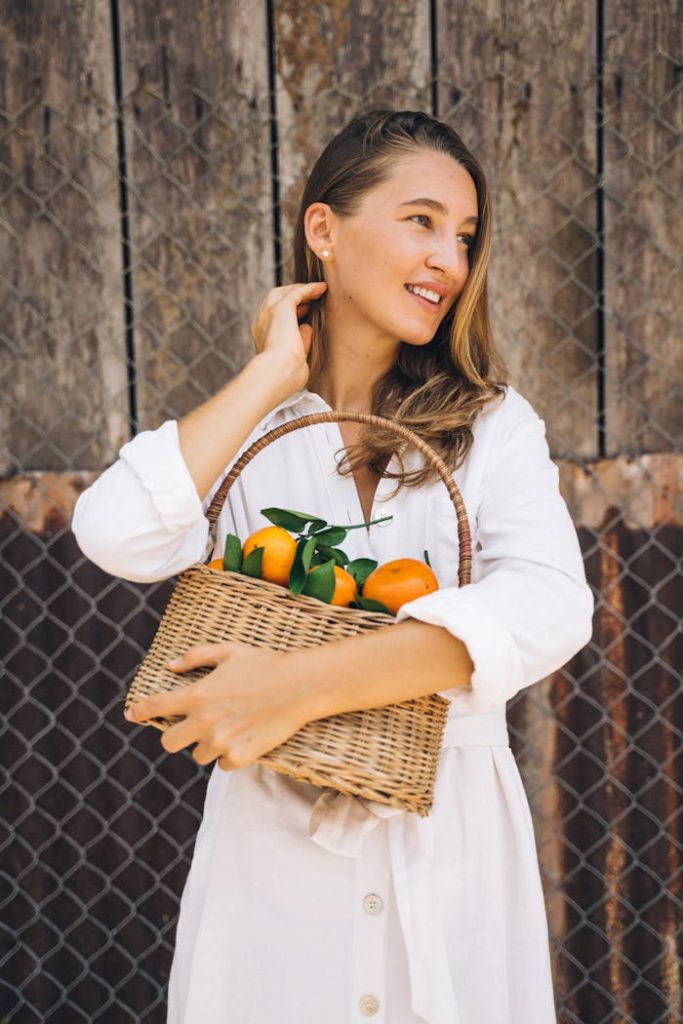 pexels-photo-7299653 Smiling woman holding a wicker basket filled with fresh oranges, wearing a white dress outdoors.